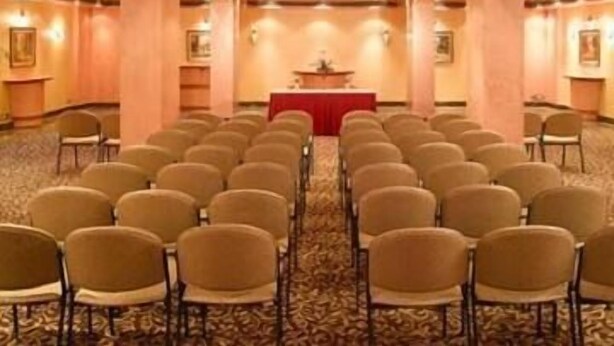 Una sala conferenze con file di sedie rivolte a un podio, pavimento in moquette e illuminazione a parete.