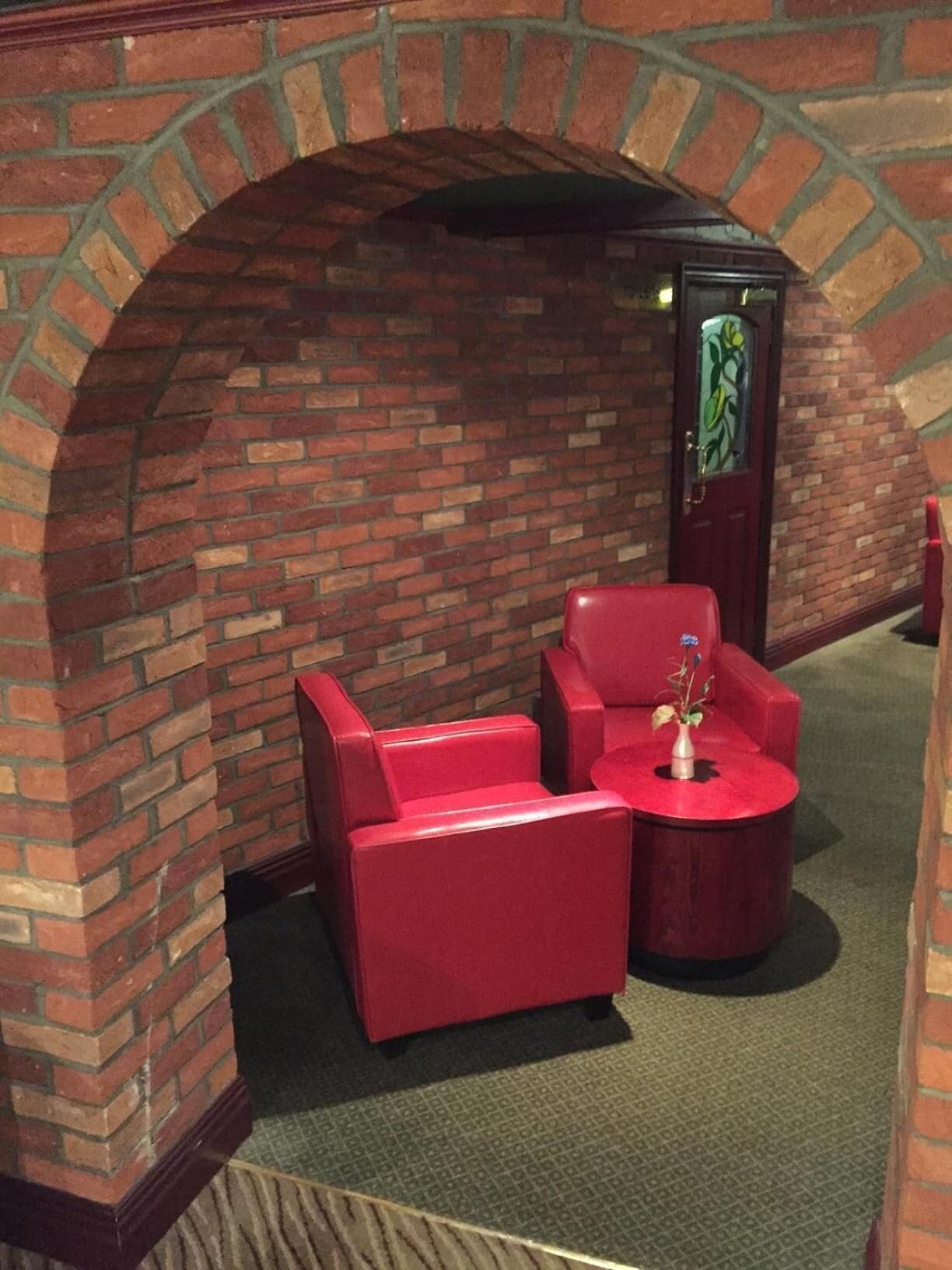 Een kamer met een bakstenen boog, rode bekleden stoelen en een kleine ronde tafel met een vaas.
