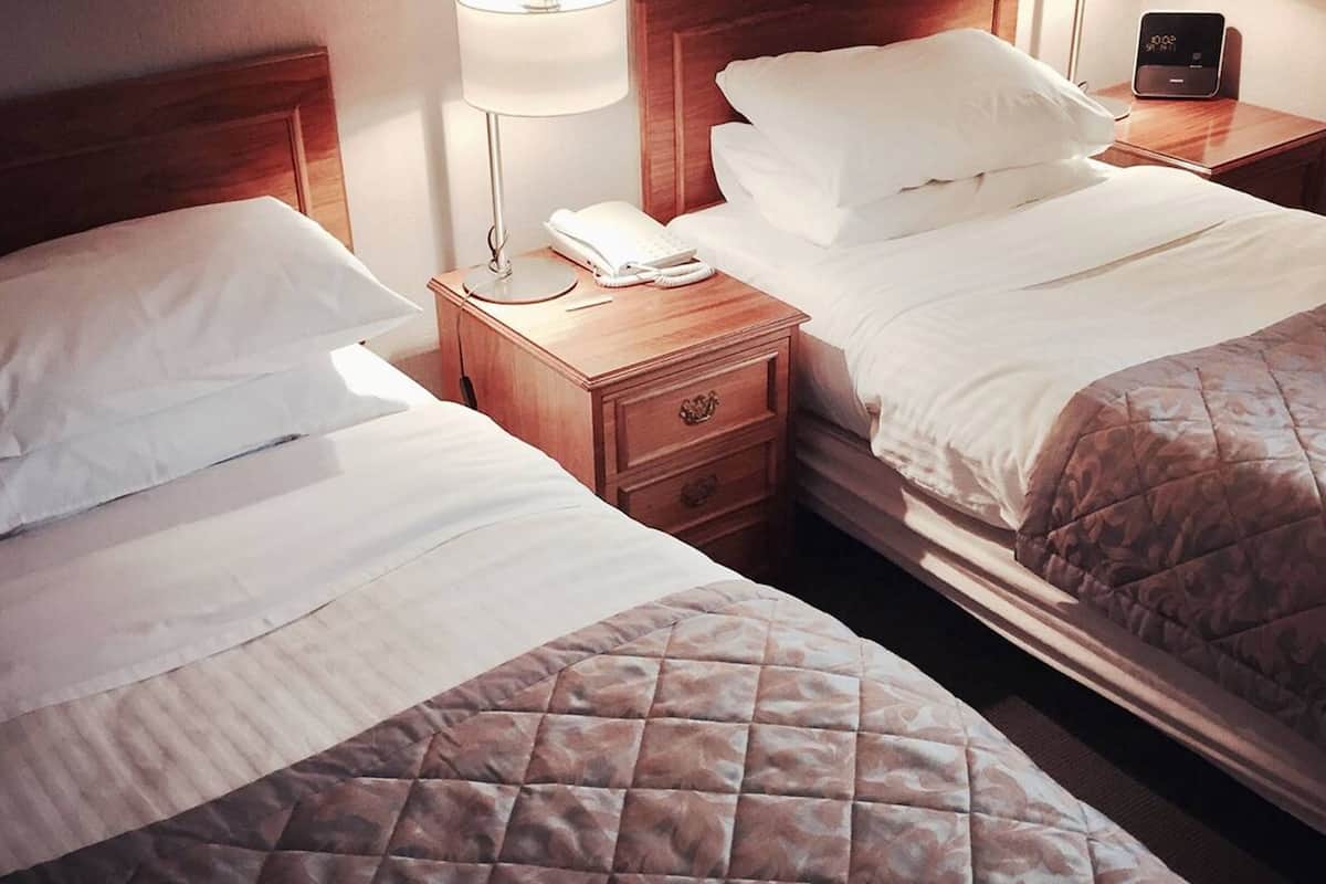 A hotel room with two beds, each with white linens and a quilted blanket. There is a bedside table with a lamp, a phone, and a digital clock.