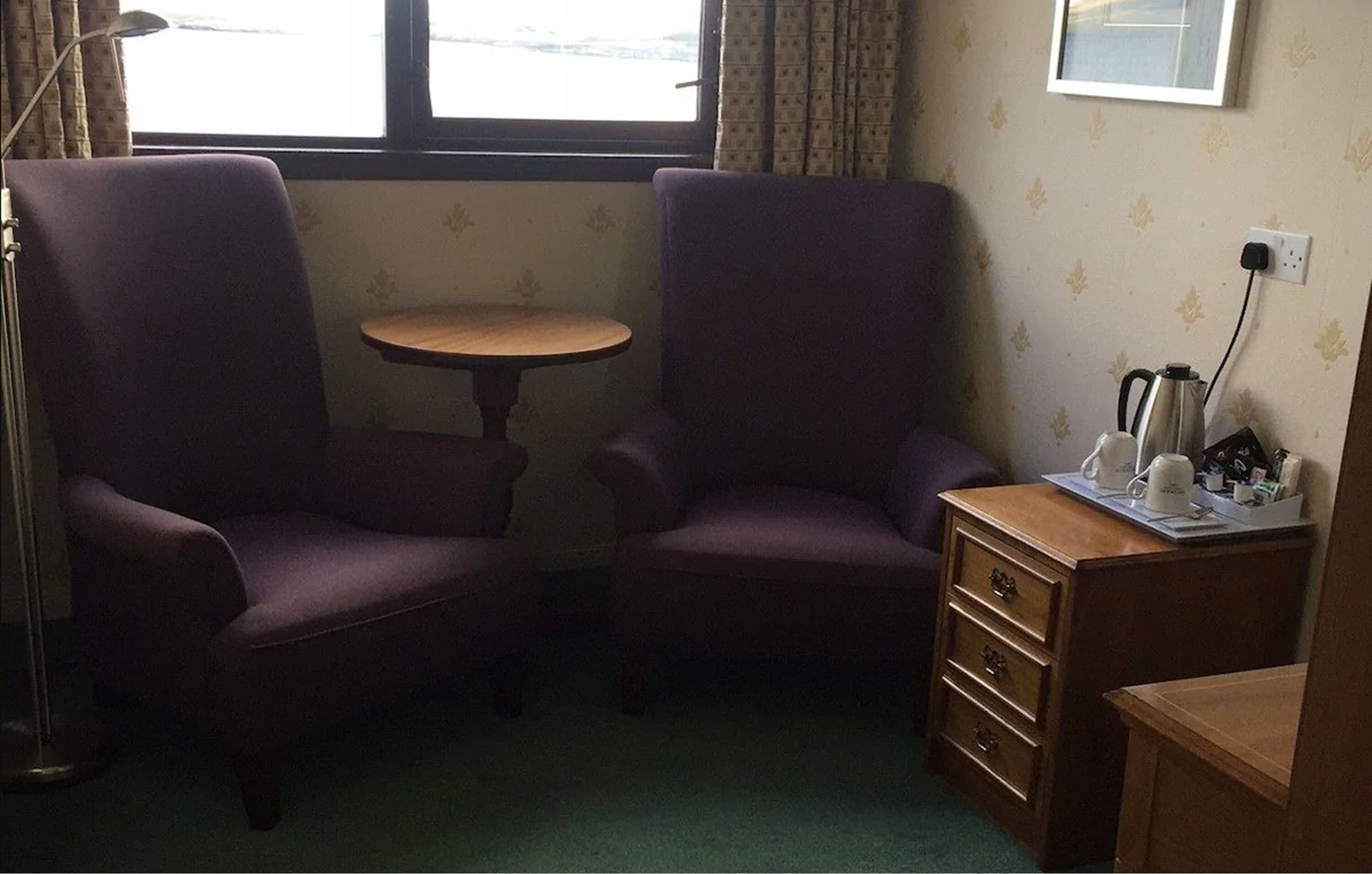 A room with two purple armchairs, a small round table, a wooden dresser, and a window with curtains.