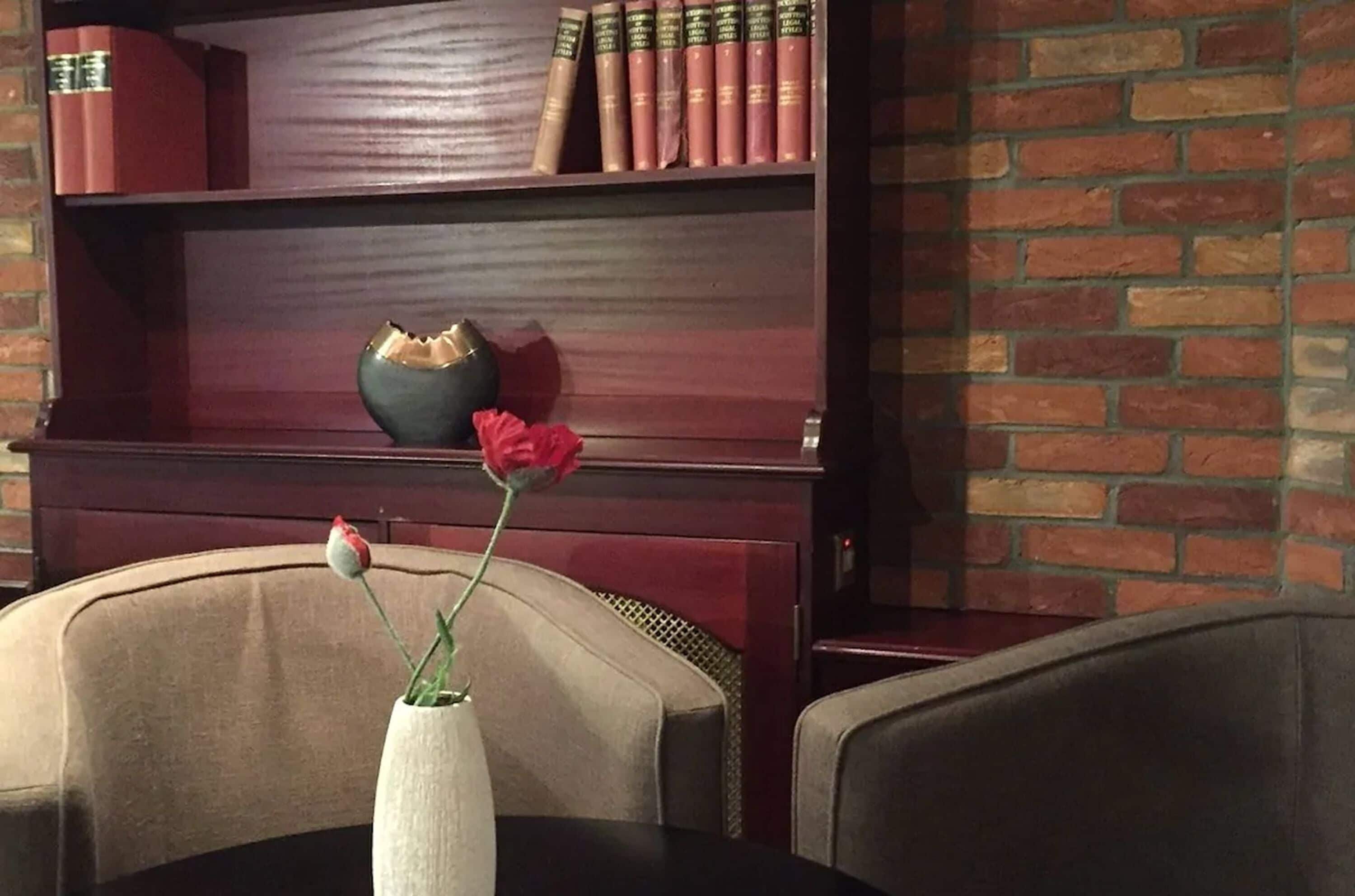 Ein Zimmer mit Backsteinwand, einem Bücherregal mit Büchern, einer Vase mit einer einzelnen Blume und zwei Stühlen.