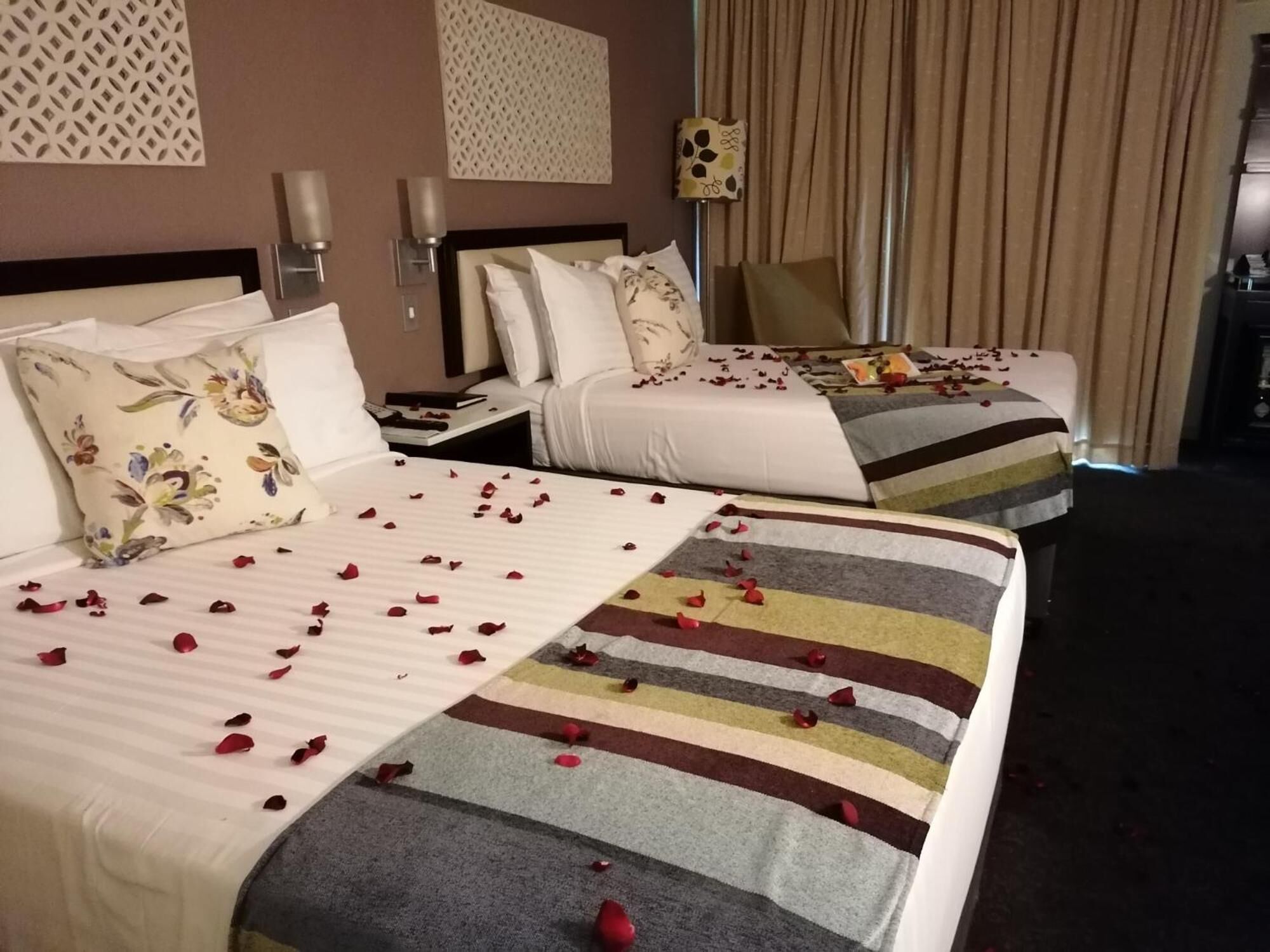Une chambre d’hôtel avec deux lits, chacun ayant un style de literie différent. On y trouve des coussins décoratifs et une table de chevet avec une lampe. La chambre est dotée d’un mur à motifs et d’un rideau.