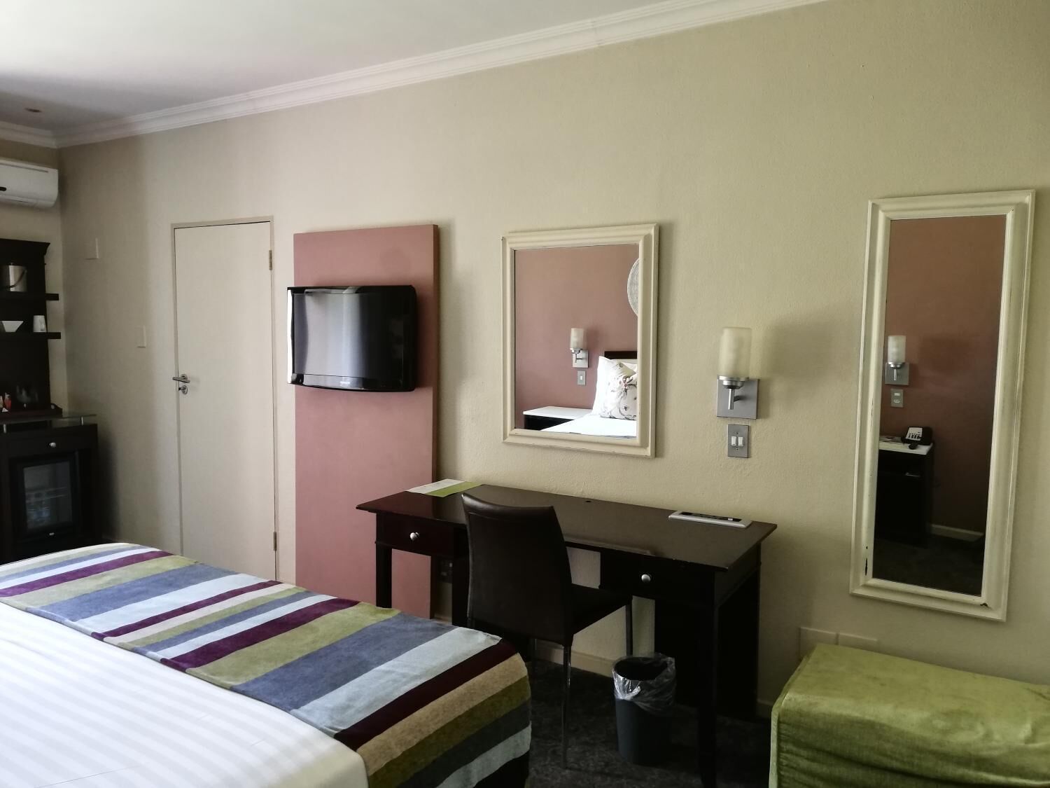 A hotel room with a bed, a desk, a chair, a television, a mirror, and a trash bin.