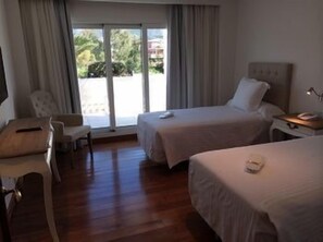 Hotelkamer met twee bedden, een bureau, een stoel en een groot raam met uitzicht op groen en gebouwen.
