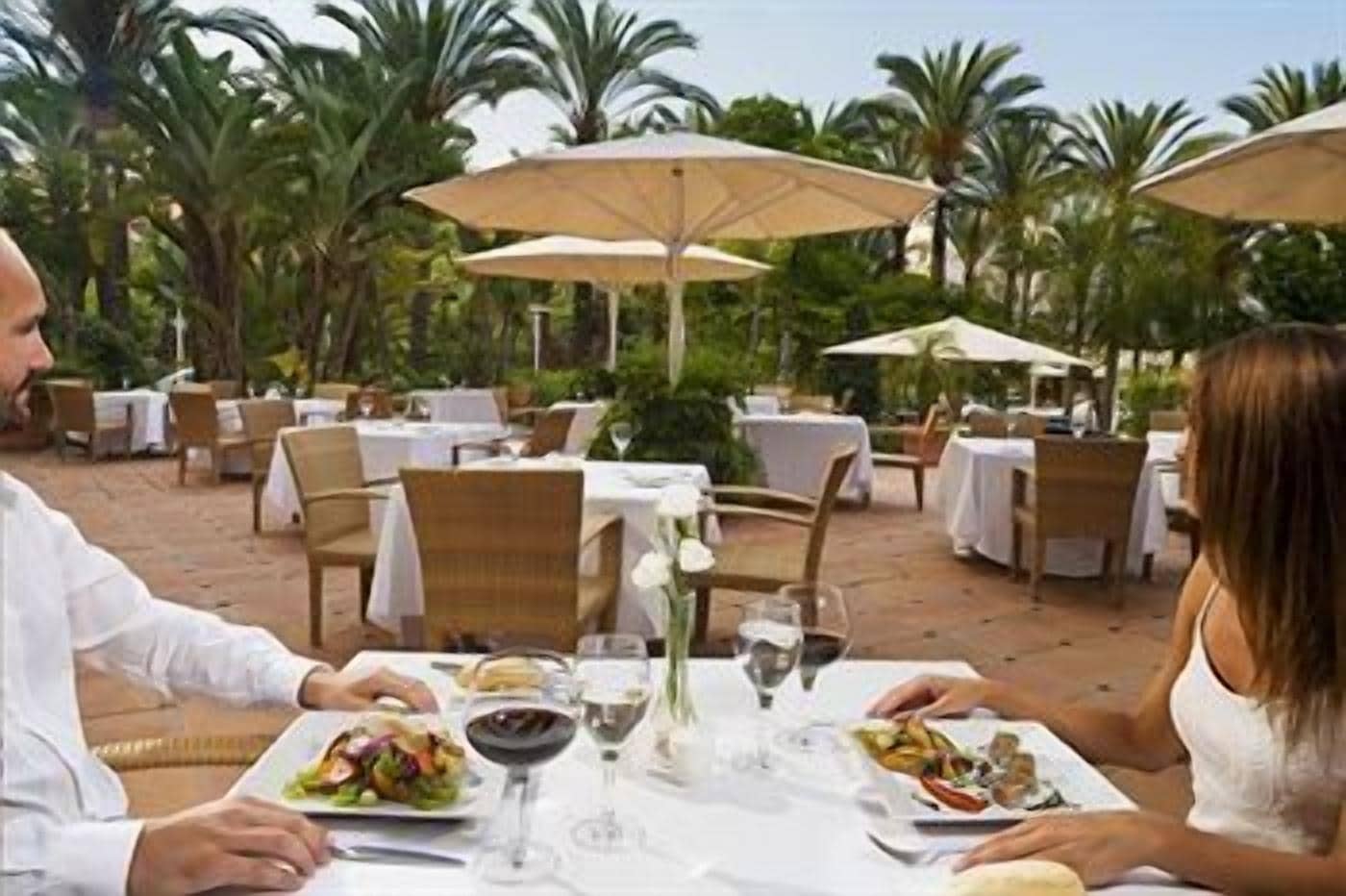 Un espacio de comedor al aire libre con mesas y sillas, sombrillas y un paisaje tropical.