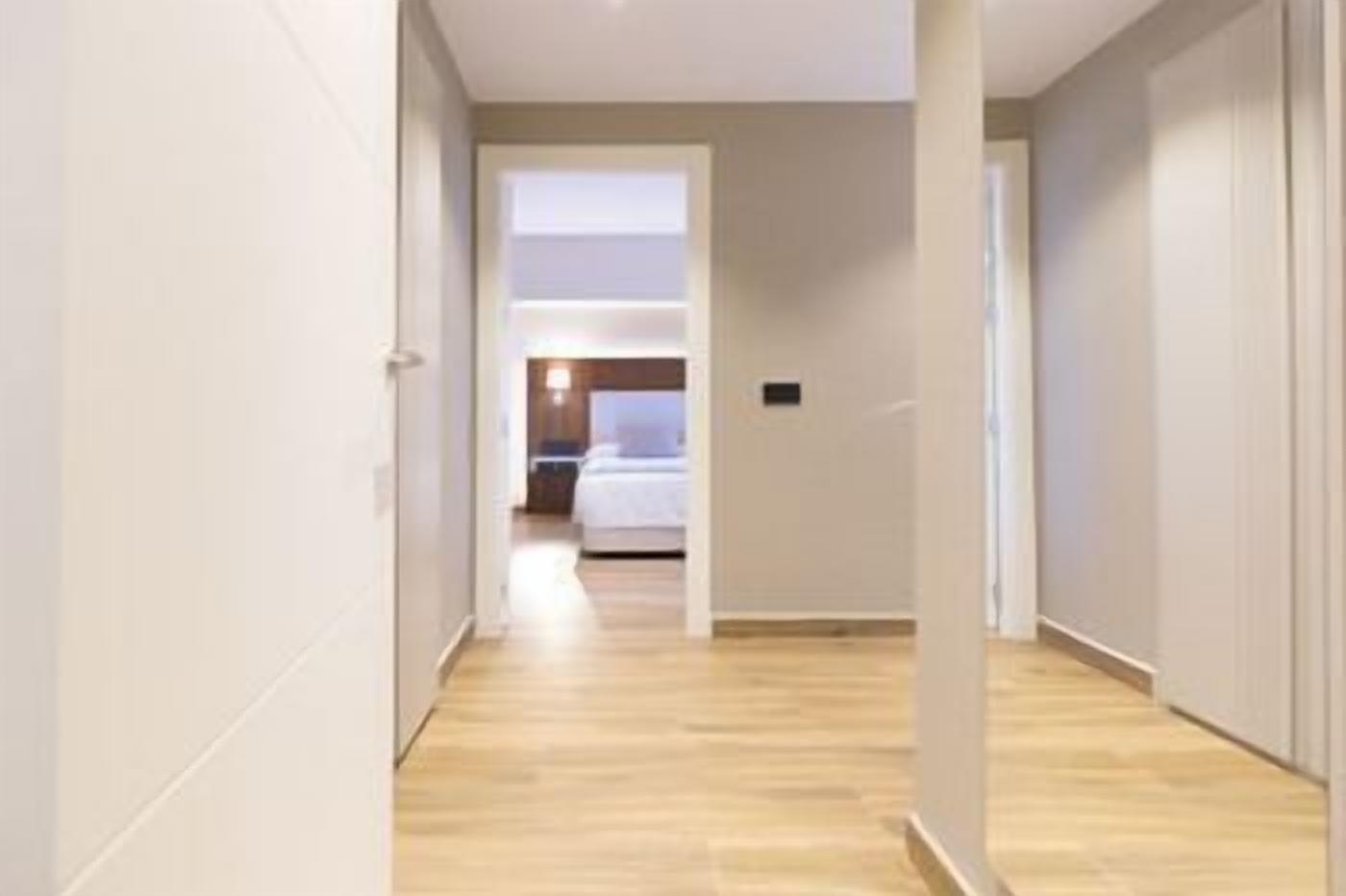 Un pasillo que conduce a un dormitorio con una cama, una mesita de noche y una lámpara fijada a la pared.