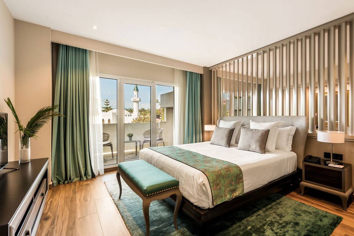 A modern hotel room with a large bed, a bench, a desk, and a view of a building with a tower.