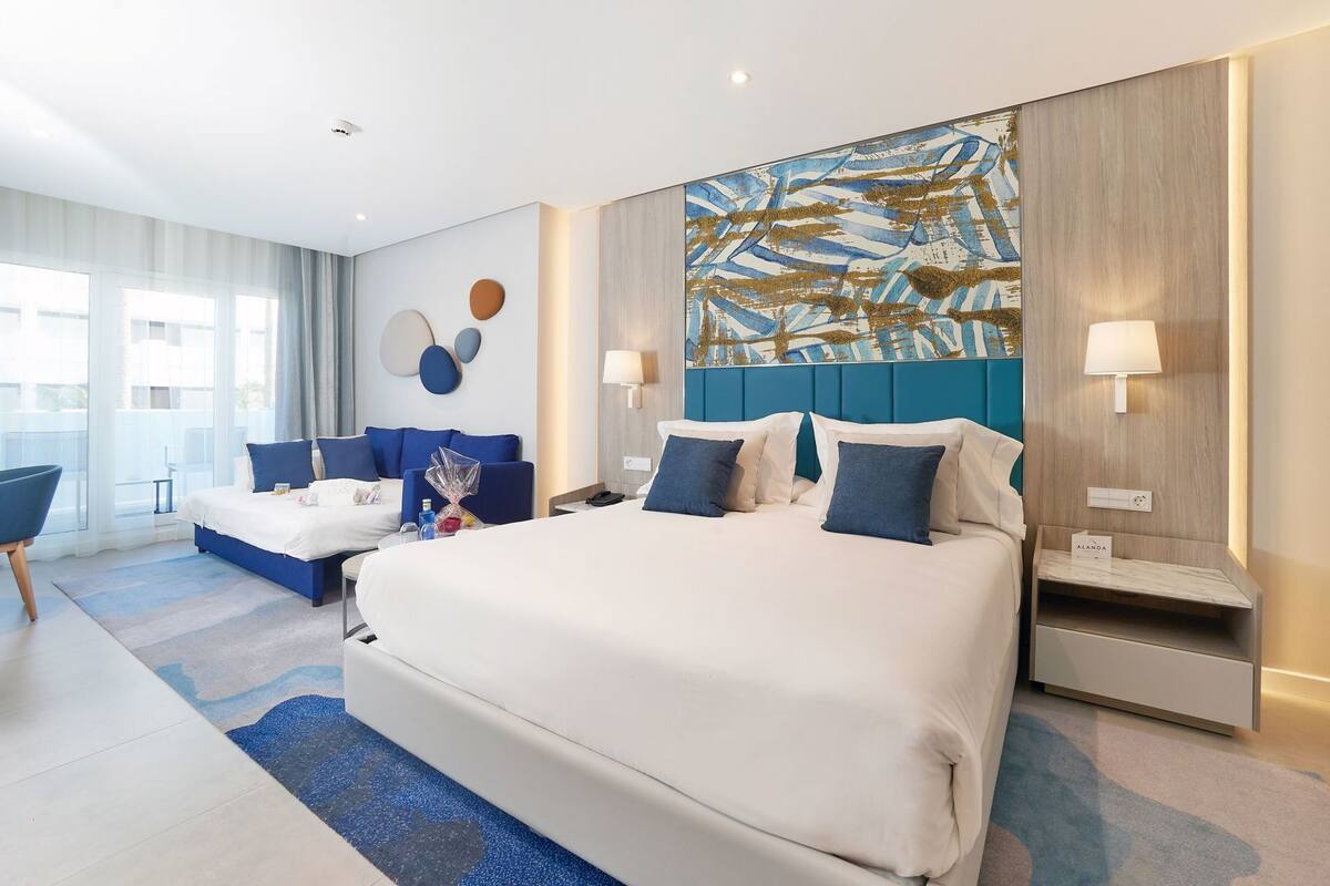 A modern hotel room with a large bed, a sofa, a desk, and a view of the city.