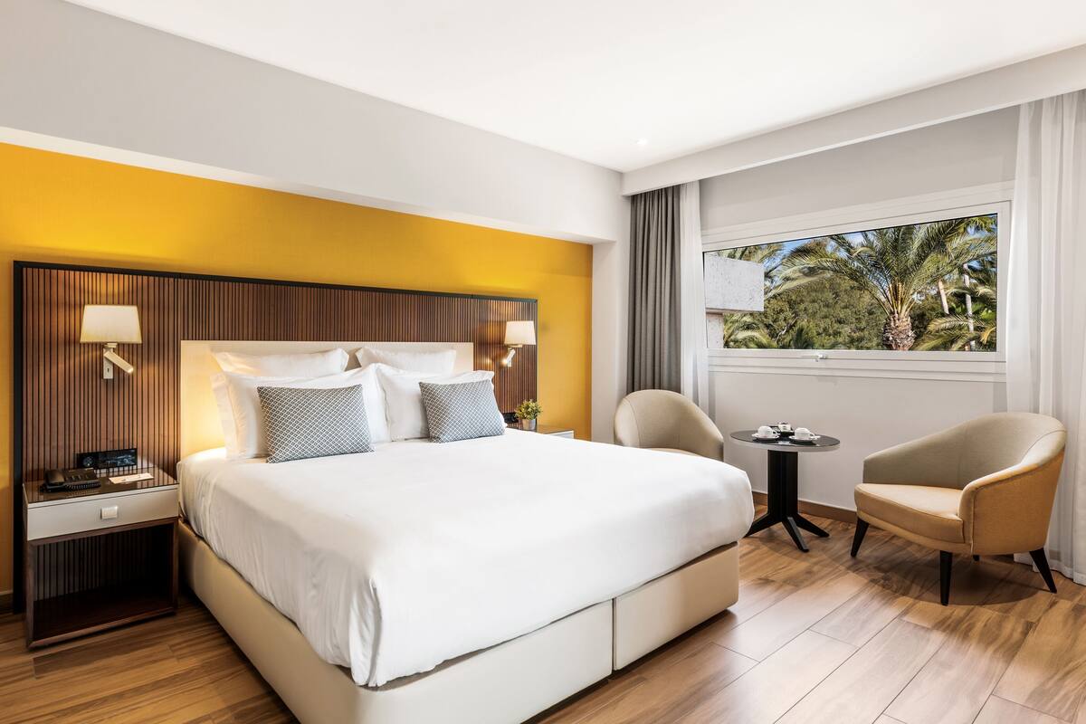 A hotel room with a large bed, bedside tables, a chair, a small table with tea cups, and a window with a view of palm trees.