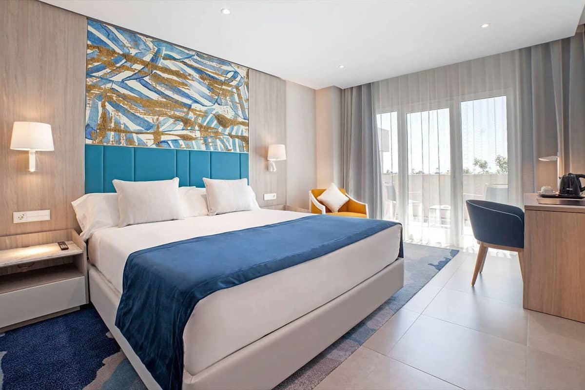 A modern hotel room with a large bed, a desk, a chair, and a view of the outdoors.
