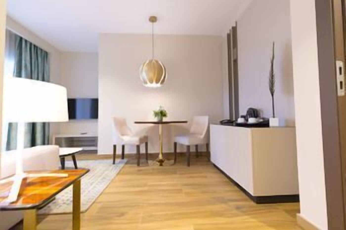 Una habitación de hotel moderna con zona de comedor, sofá, mesa y lámpara colgante.