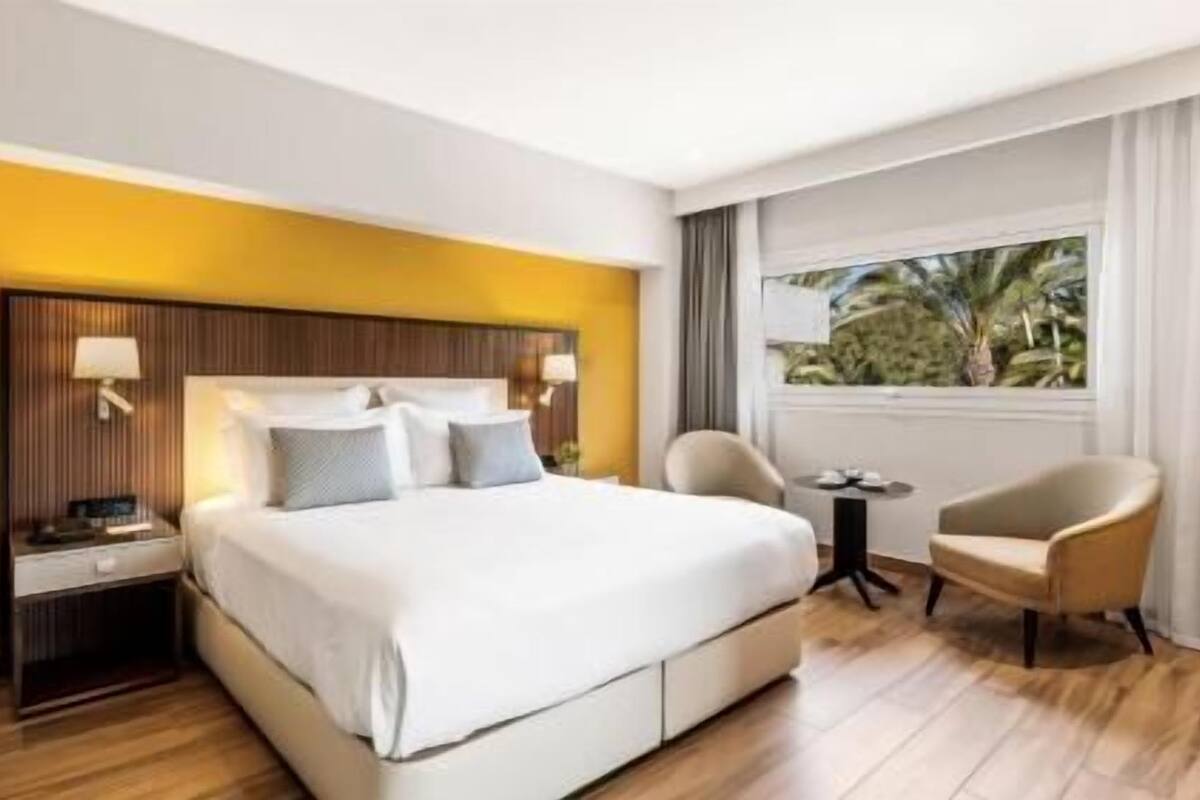 A hotel room with a bed, bedside tables, a chair, a small table, and a window with a view.
