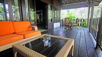A covered outdoor area with a wicker sofa and a glass-top table, wooden flooring, and a view of the surrounding greenery.
