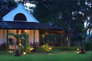 Un edificio con techo triangular, un patio cubierto y un jardín bien cuidado con palmeras y plantas.