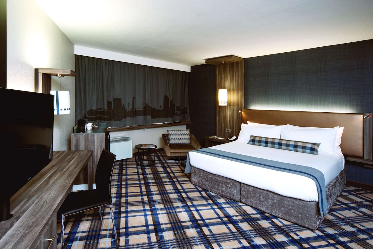 A hotel room with a large bed, a desk, a chair, a television, and a cityscape view.