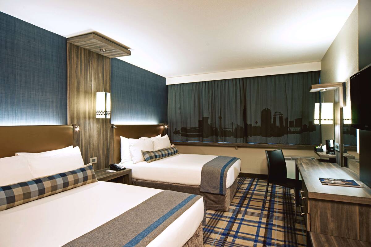 A hotel room with two beds, a desk, a chair, and a large window with curtains.
