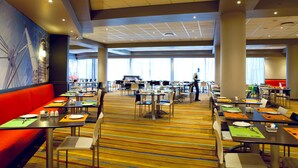 A spacious dining area with multiple tables and chairs, a large mural on the wall, and a view of the city through large windows. - Garden Court Milpark (Johannesburg)