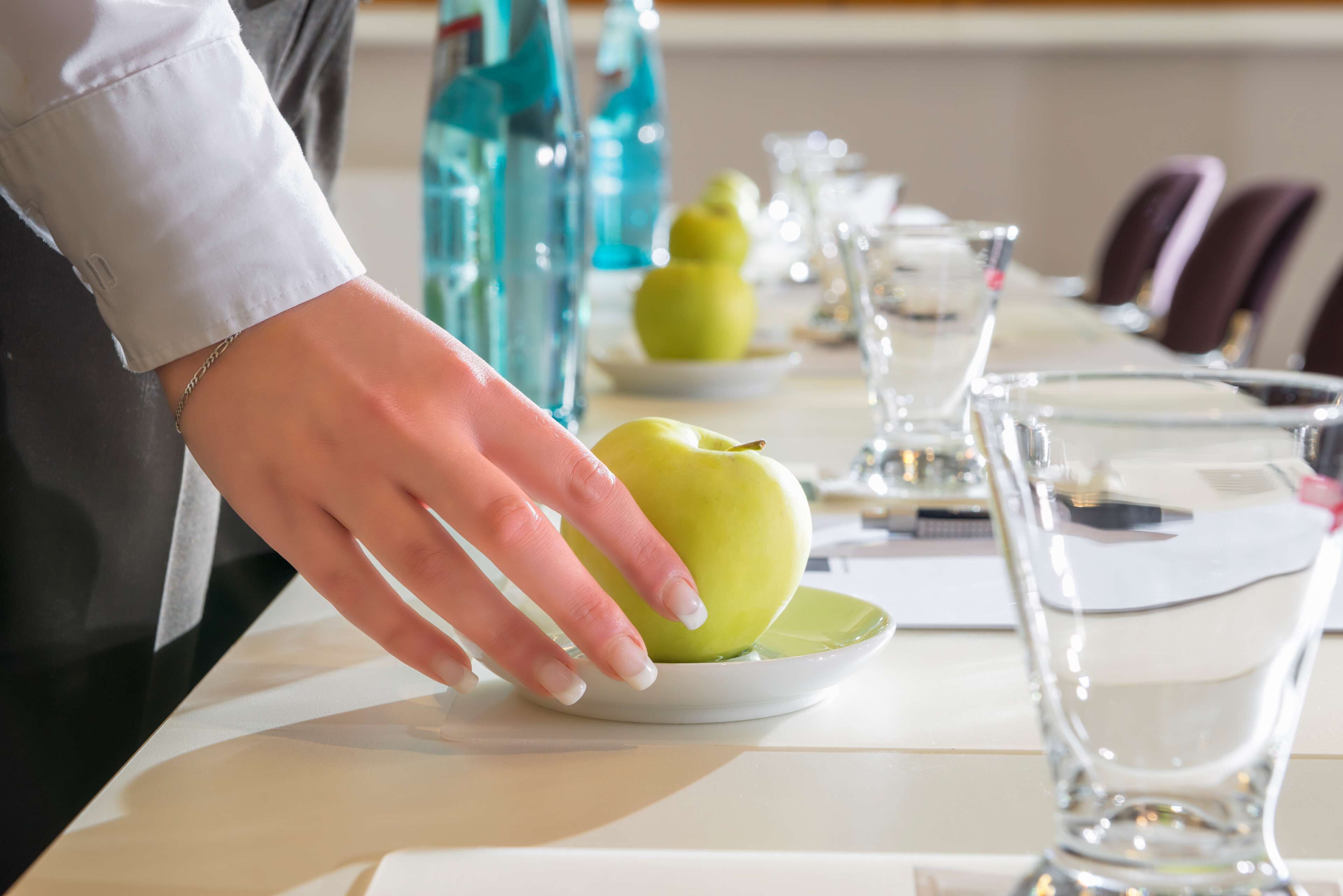 A hand placing a green apple on a white plate on a table with glasses and water bottles.