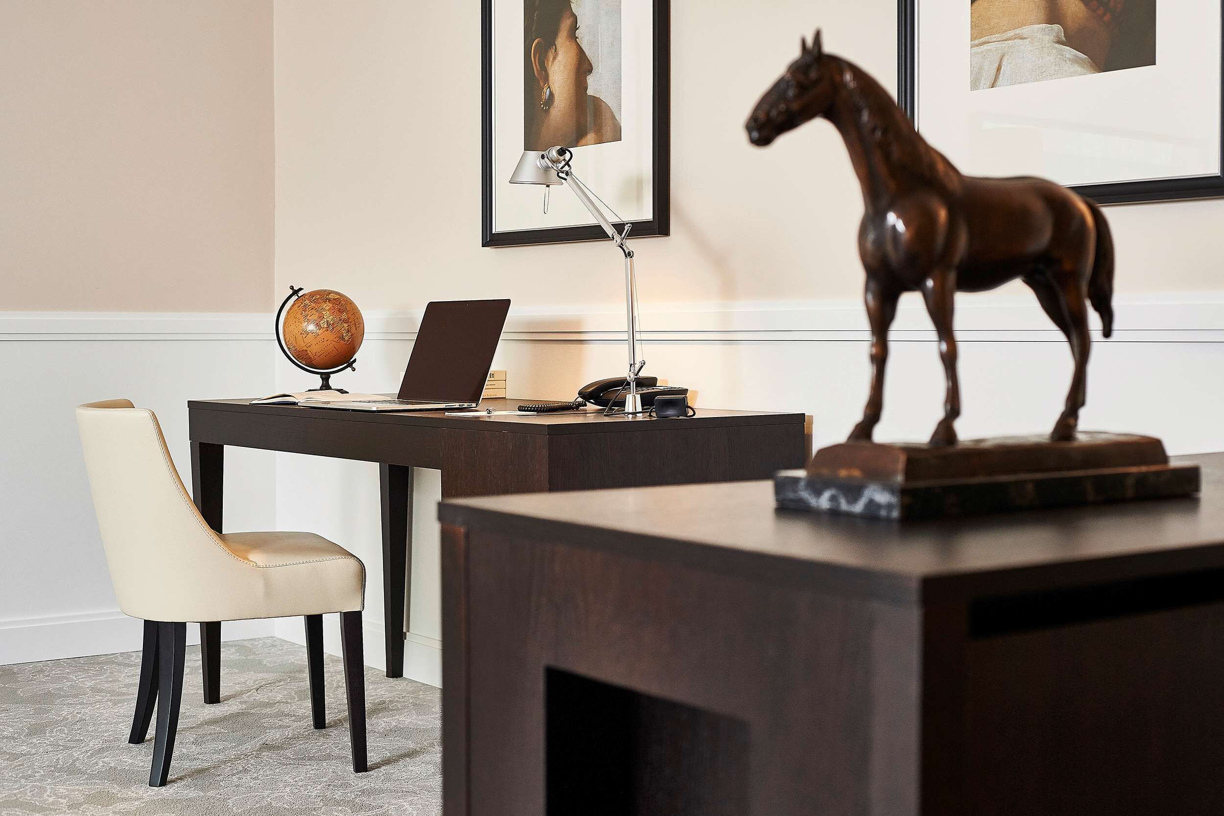 A desk with a laptop, a globe, and a telephone. There is a chair and a wooden horse sculpture.