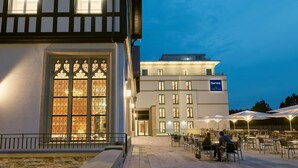 L’extérieur d’un hôtel, avec un bâtiment de style traditionnel et une aile moderne, un espace extérieur aménagé pour s’asseoir et une entrée bien visible.