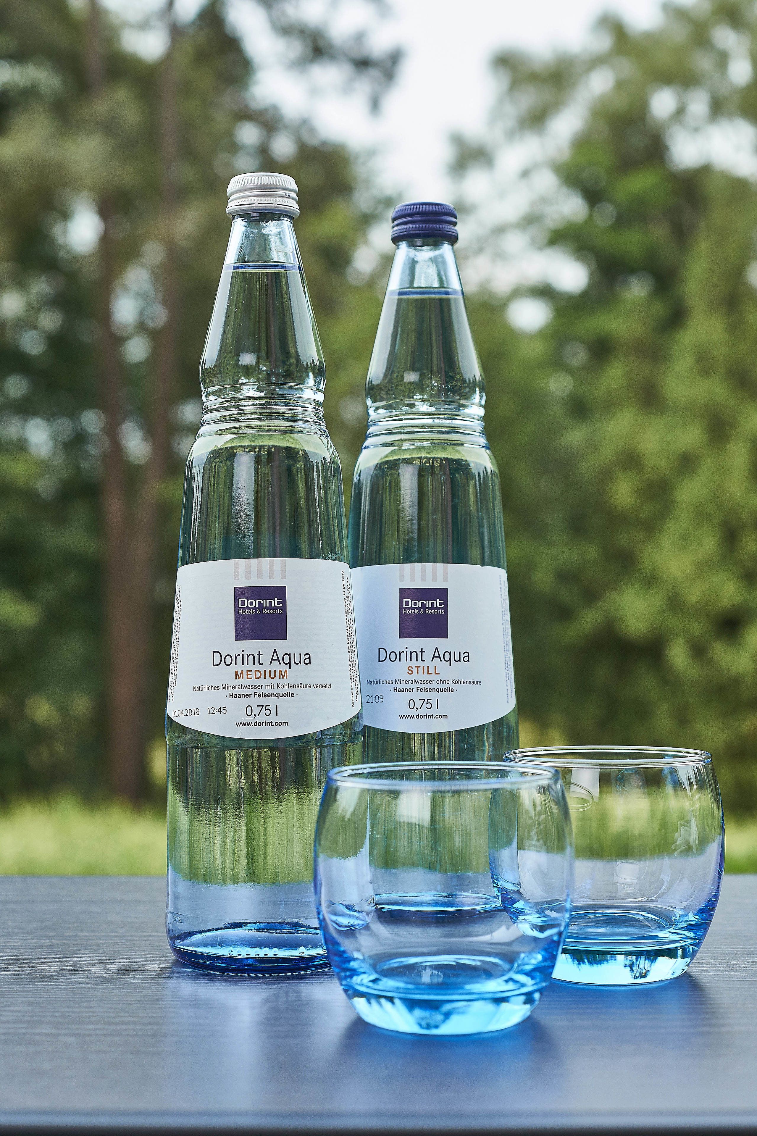 Zwei Flaschen Dorint Aqua Medium und zwei Gläser auf einem Tisch vor unscharfem, grünem Hintergrund.