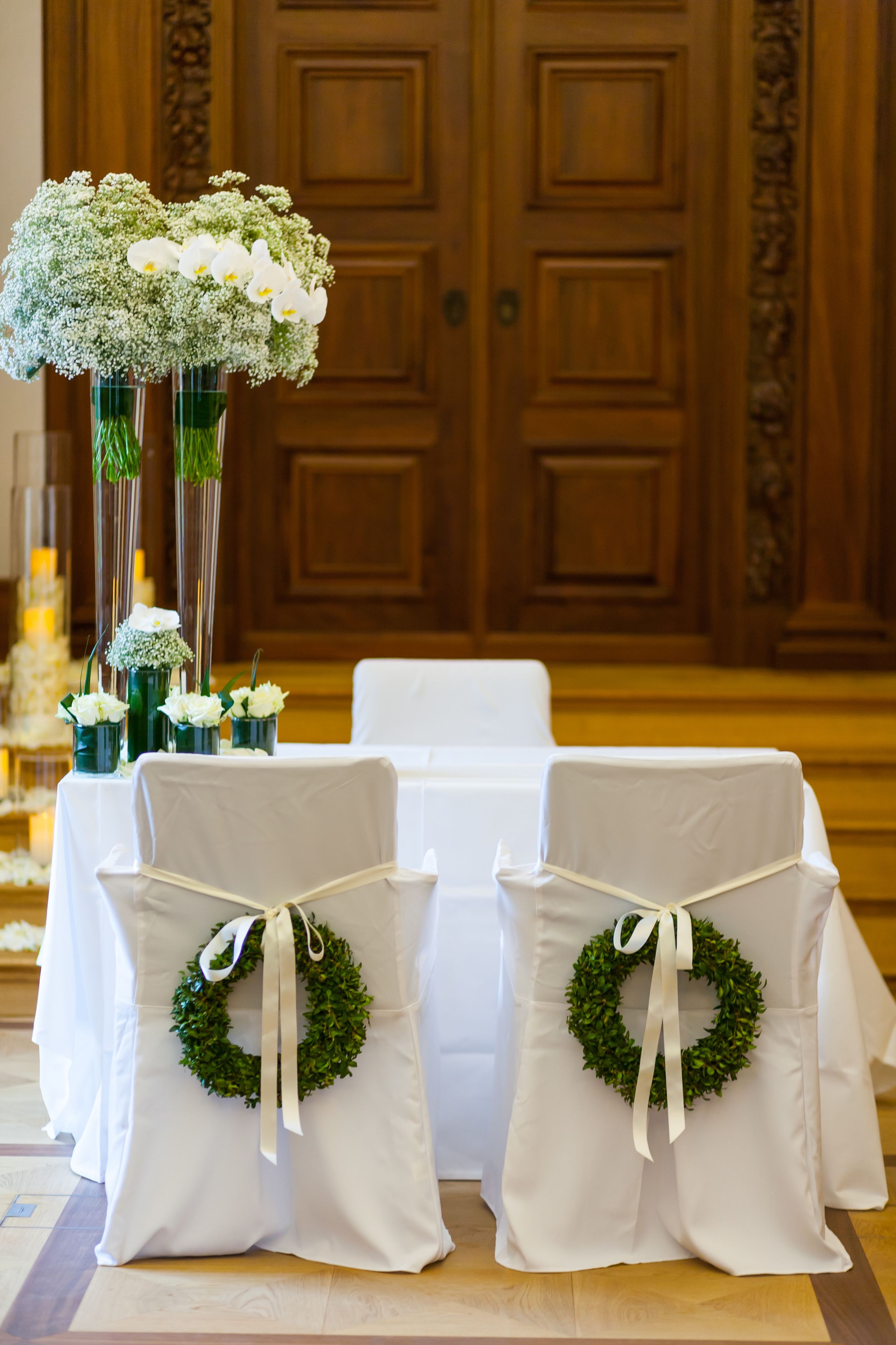 Un área de recepción con una puerta de madera, sillas blancas adornadas con coronas verdes y una mesa con manteles blancos.