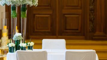 Un área de recepción con una puerta de madera, sillas blancas adornadas con coronas verdes y una mesa con manteles blancos.