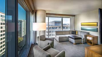 A modern hotel room with a large window, a sofa set, a lamp, and a small wooden side table.