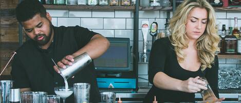 A bar setting with two bartenders preparing drinks. There are various bottles and jars of ingredients on the counter.