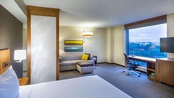 A hotel room with a bed, a desk, a chair, a sofa, and a large window offering a city view.
