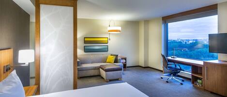 A hotel room with a bed, a desk, a chair, a sofa, and a large window offering a city view.