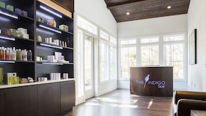 Een ontvangstbalie van een spa met een houten vloer, grote ramen en een receptiebalie met de tekst 'The Indigo Spa'.