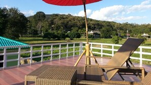 Un balcon agrémenté de fauteuils en osier et d’un parasol rouge, offrant une vue sur un paysage verdoyant avec des collines et des maisons.