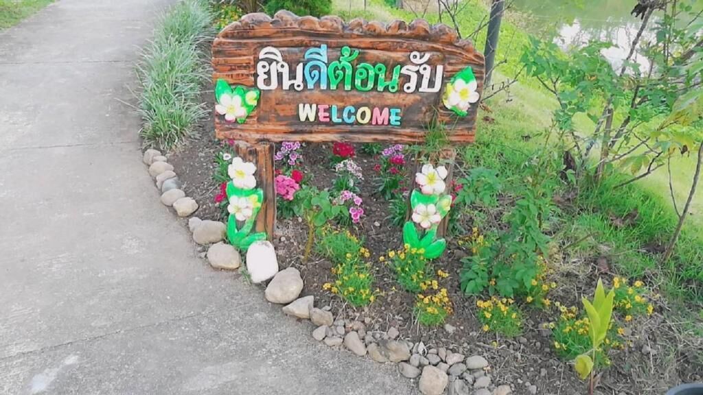 A wooden welcome sign with Thai script and English text, surrounded by flowers and plants.