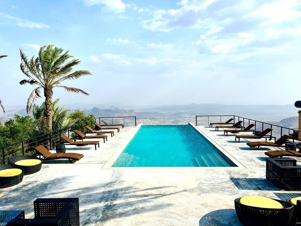 A rooftop infinity pool with lounge chairs and a palm tree, overlooking a cityscape. - The View (Al Hamra)