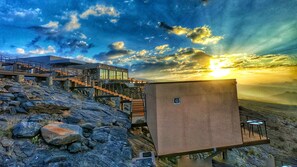 A modern building with a staircase leading to a viewing platform, overlooking a rocky landscape and a sunset.