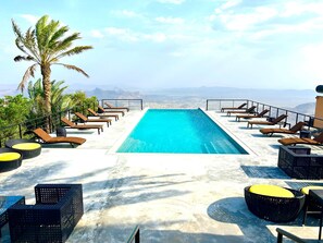 A rooftop infinity pool with lounge chairs and a palm tree, overlooking a mountainous landscape. - The View (Al Hamra)