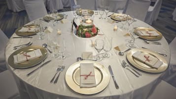 A round dining table set for a formal event with white tablecloths, gold-rimmed plates, and silver cutlery.