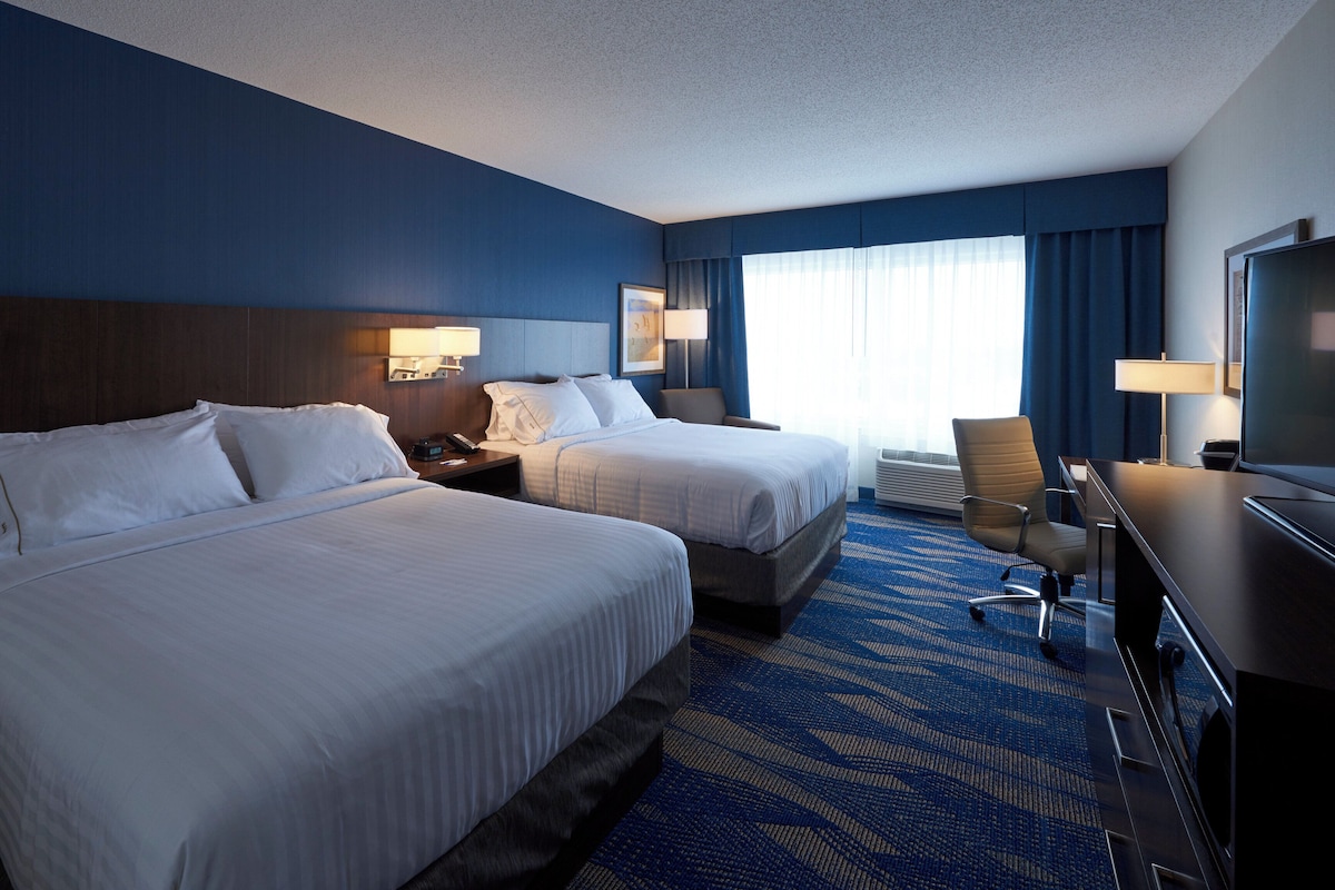 A hotel room with two beds, a desk, a chair, a TV, and a window with curtains.