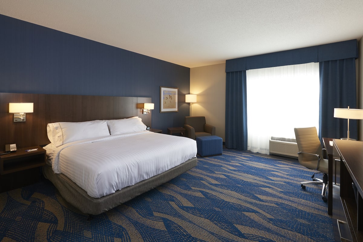 A hotel room with a large bed, a desk, a chair, a desk lamp, a wall-mounted phone, and a window with curtains.