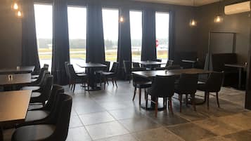 A restaurant with multiple tables and chairs, large windows with curtains, and hanging light fixtures.