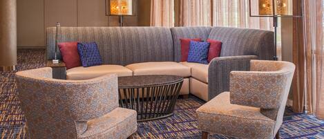 A modern hotel lobby with a sectional sofa, two armchairs, a coffee table, and a patterned carpet.
