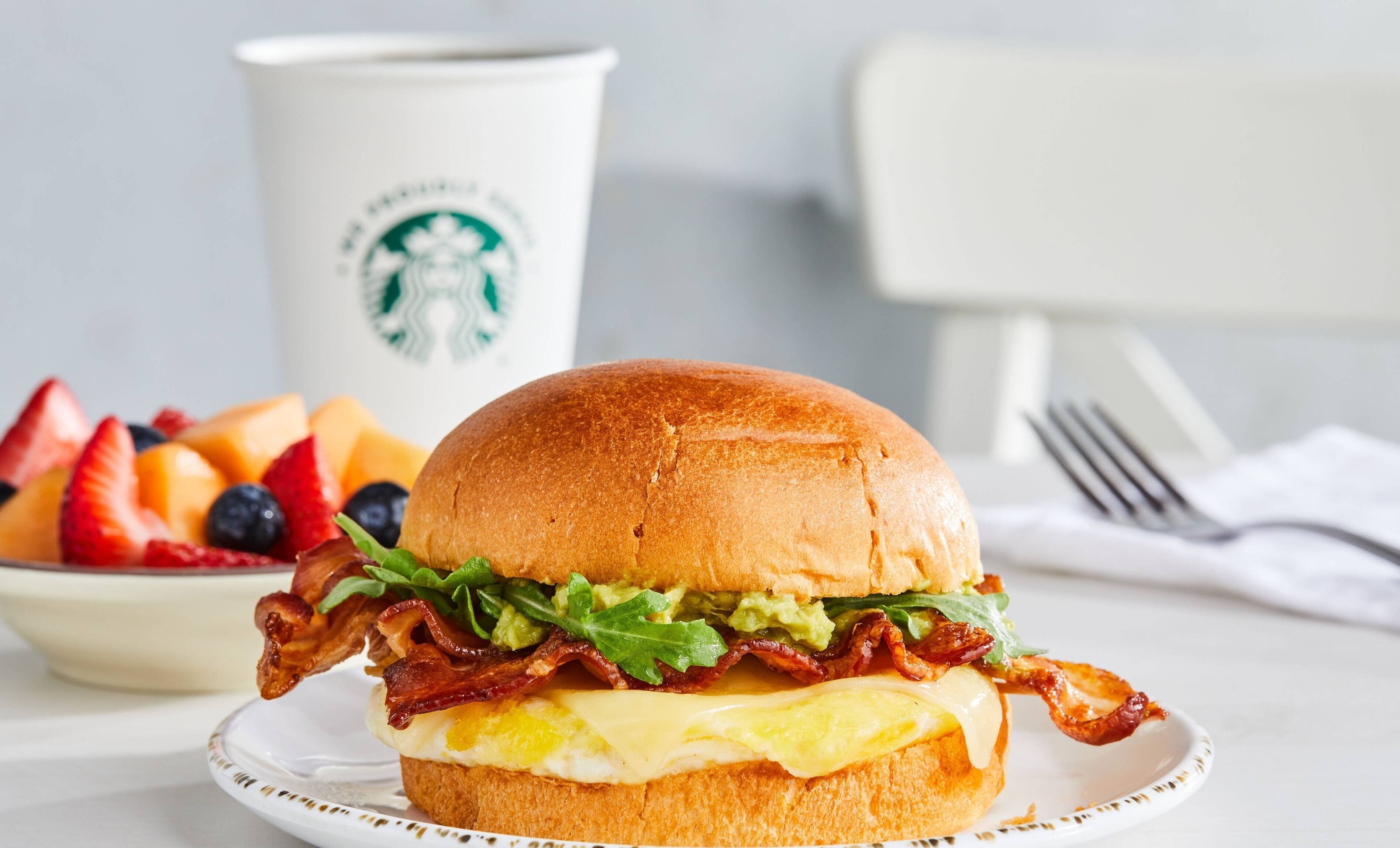 A breakfast sandwich with bacon, cheese, and lettuce on a bun, a plate of fruit, and a Starbucks cup in the background.