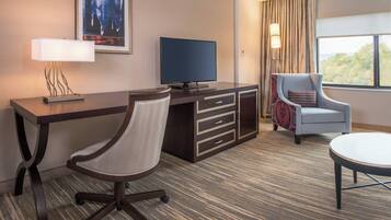 A hotel room with a desk, a chair, a television, a lamp, a window with curtains, and a view of trees.