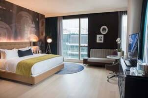 Habitación de hotel con una cama grande, un escritorio con computadora, un sofá y un ventanal con vistas a edificios.