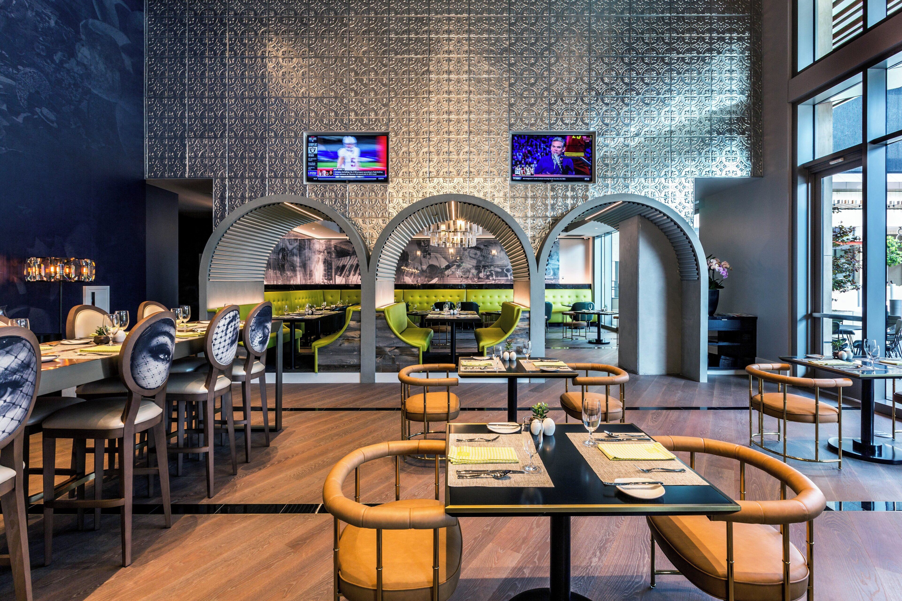 Un interior de restaurante moderno con una zona de estar central, varias mesas y dos televisores instalados en la pared.