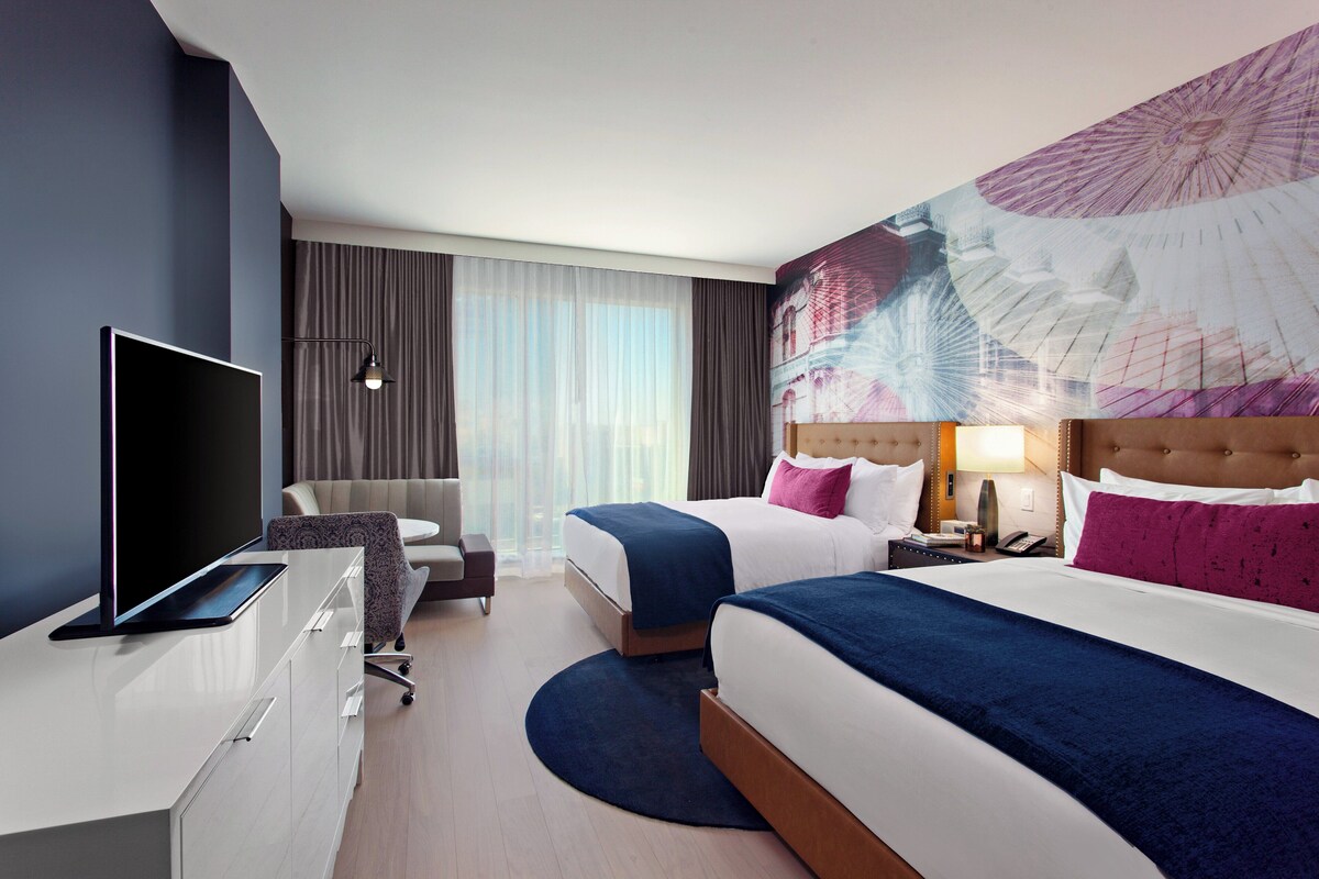 Habitación de hotel con una cama, un escritorio con televisor, una silla, una mesita de noche y un ventanal con cortinas.