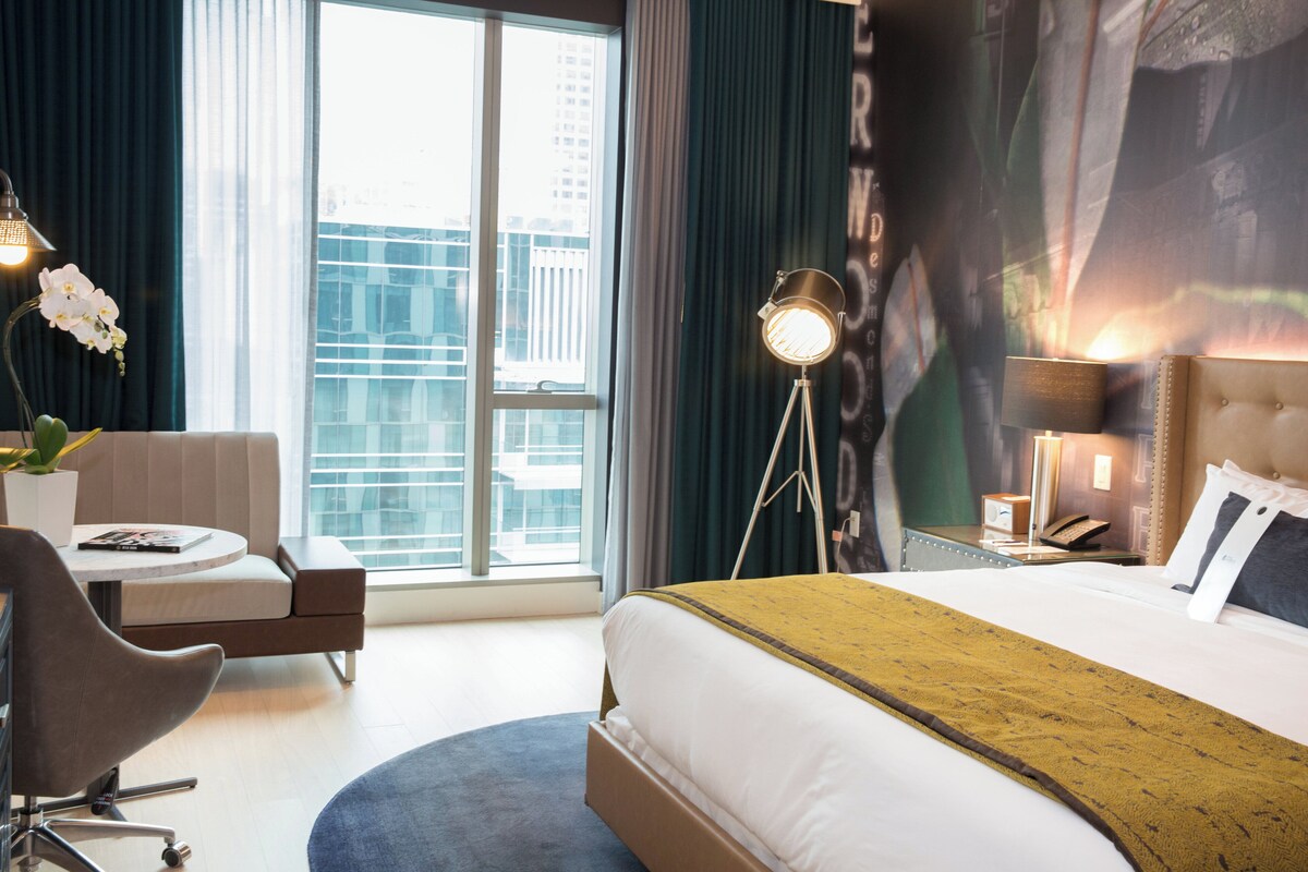 Habitación de hotel con una cama grande, un escritorio, una silla, una lámpara y vistas a la ciudad desde la ventana.