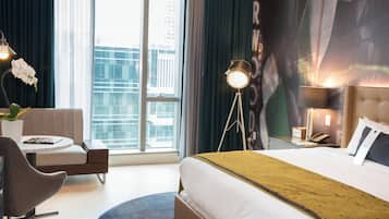 A hotel room with a large bed, a desk, a chair, a lamp, and a view of the city through the window.