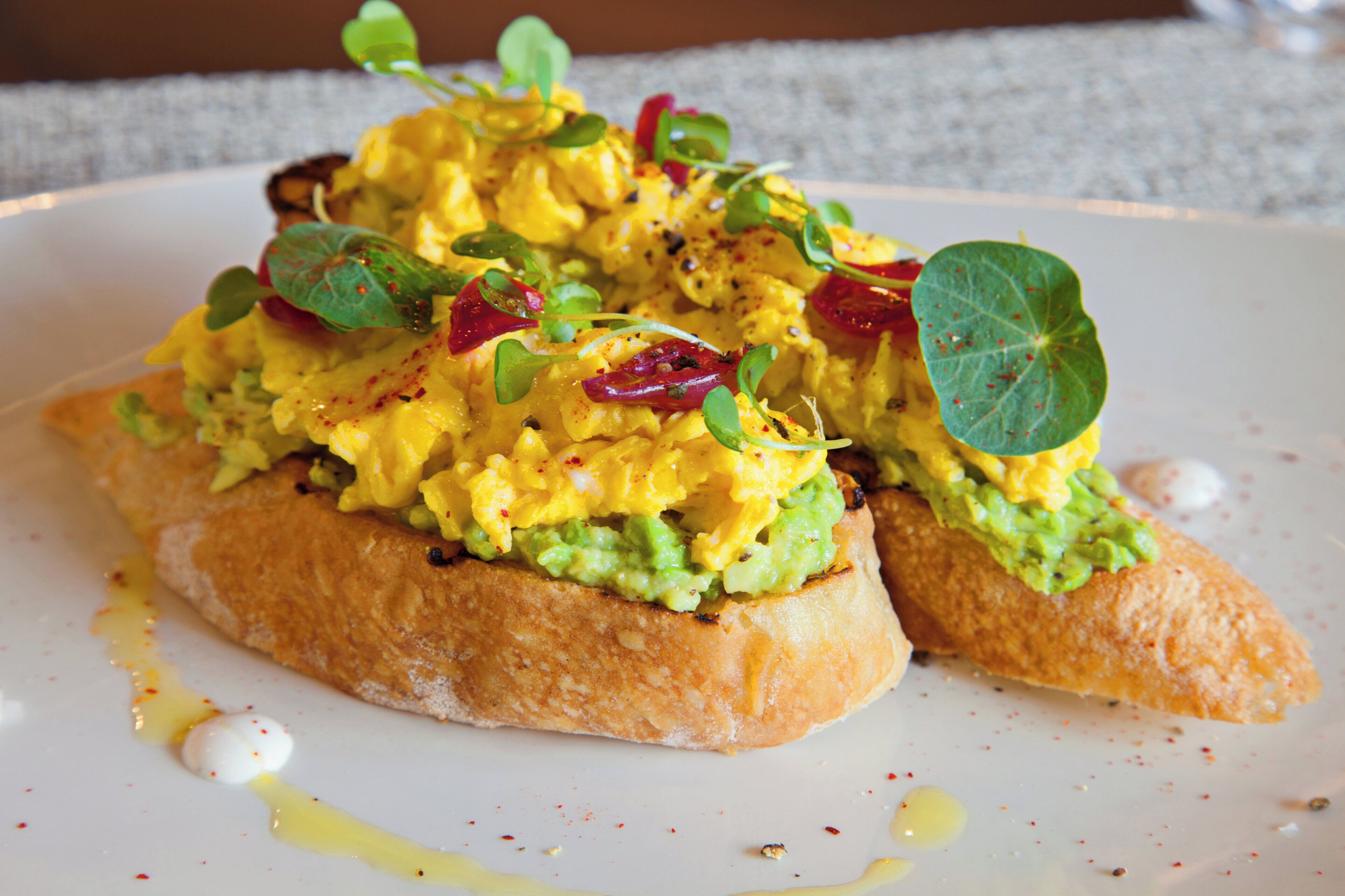 A dish with scrambled eggs, avocado, and greens on toast, served on a white plate with a drizzle of sauce.